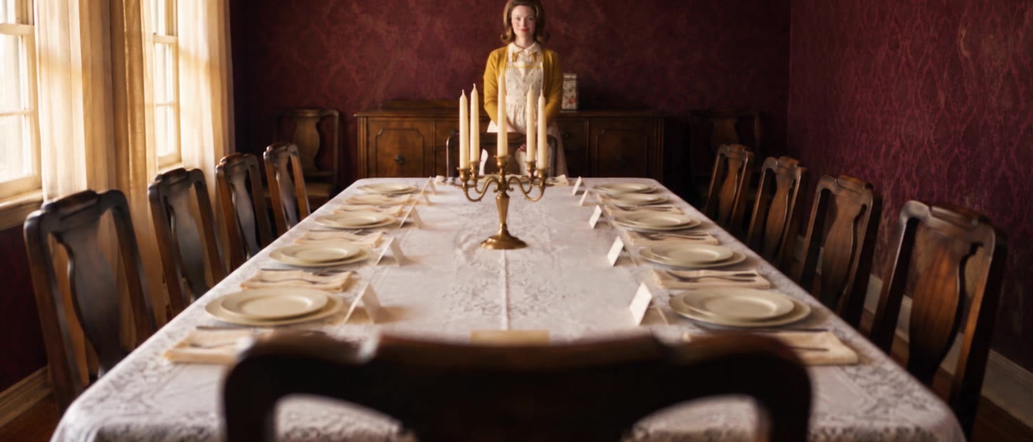 Still from Grace: a 1961 dining room, a long table set for twelve on white lace, an empty chair in the foreground.